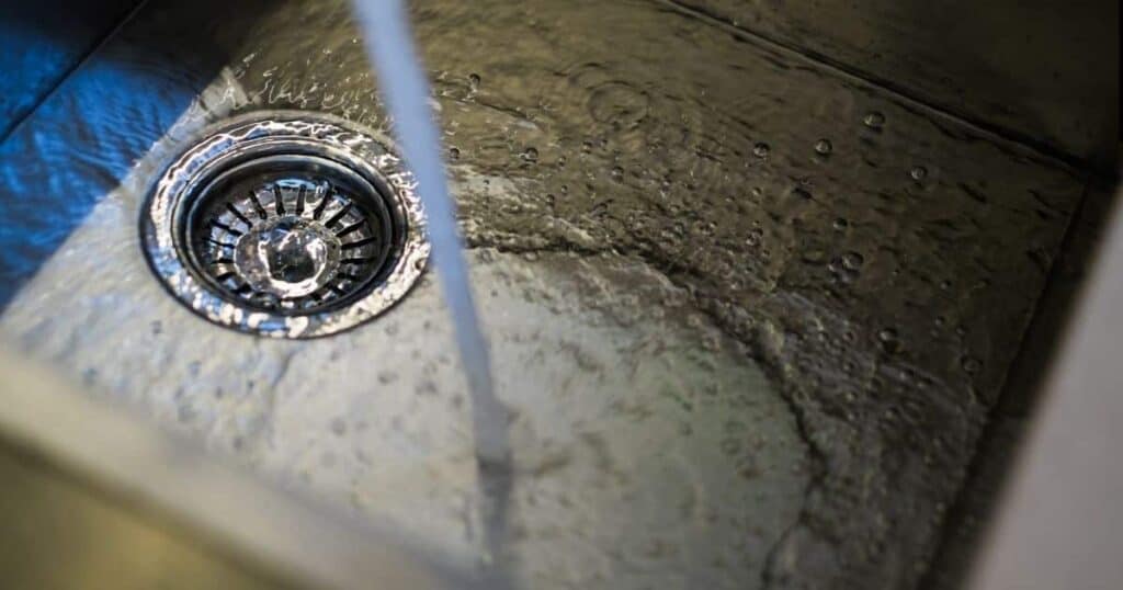 Water Is Running From A Faucet Into A Stainless Steel Sink, Creating Ripples Around The Drain.