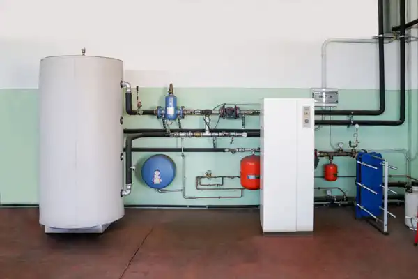 HVAC system in a utility room with pipes, a cylindrical tank, control panel, and various colored components against a two-tone wall.